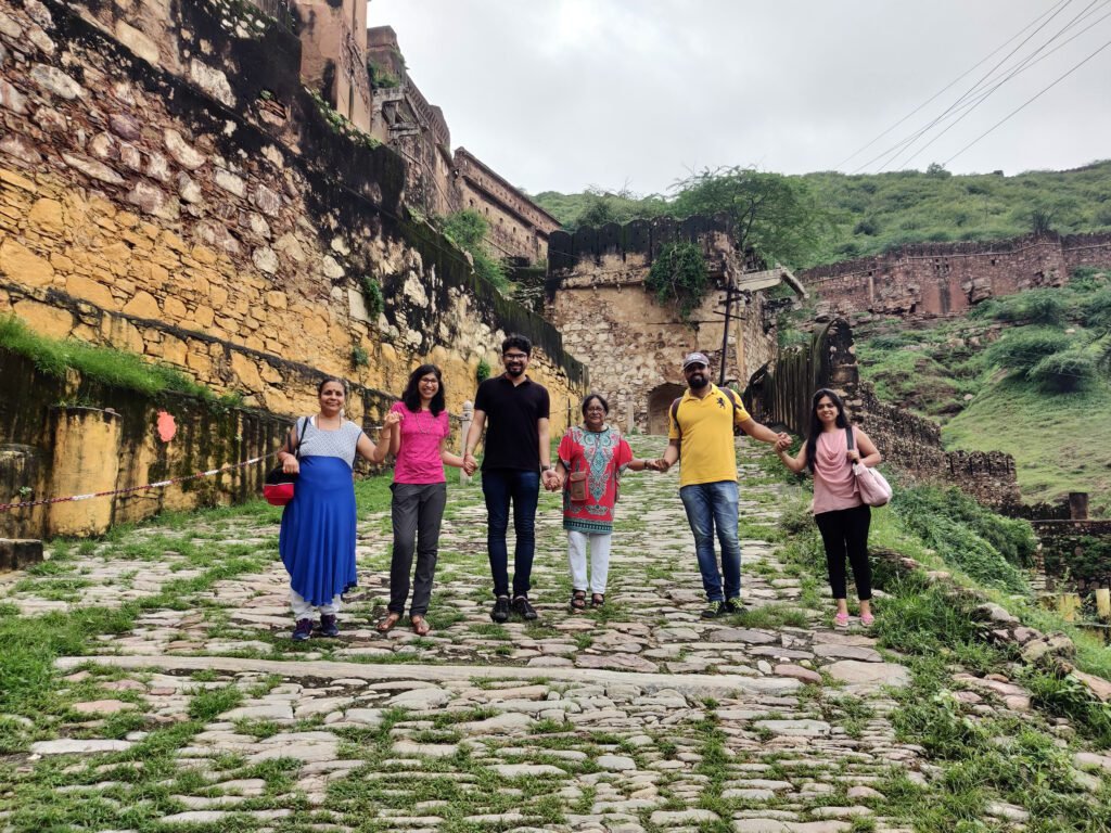 A group of six travelers, including the tour leader in a pink t-shirt, stand in a line holding hands on a cobblestone pathway leading up to the historic Taragarh Fort in Bundi. They are smiling at the camera, flanked by weathered stone fort walls on the left and a lush green hillside on the right under an overcast sky.