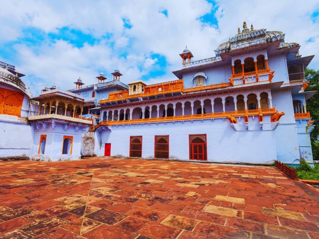 A wide-angle, eye-level view of a grand palace structure with white walls accented by bright orange borders, ornate columns, and arched balconies. The building features traditional Rajasthani architectural details, including small dome pavilions (chhatris) on the roof and elephant-shaped brackets under the upper balconies. In the foreground is a spacious, open courtyard paved with large, reddish-brown stone tiles, all set under a bright blue sky filled with scattered white clouds.