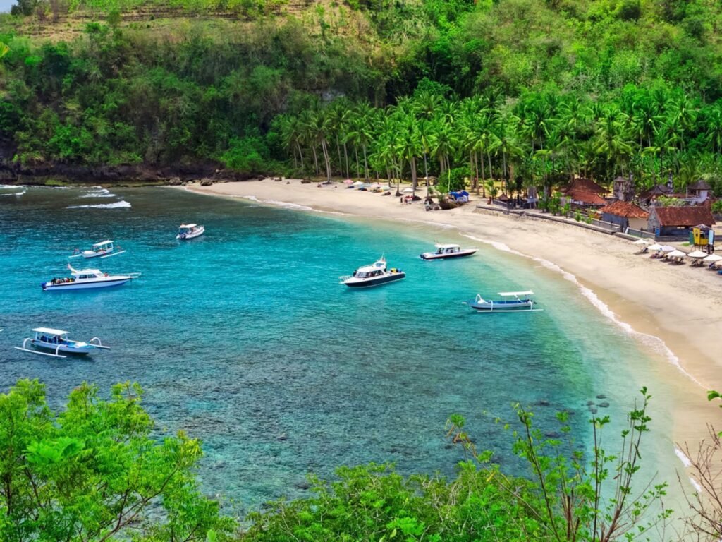 A high-angle view of Crystal Bay in Nusa Penida, showing several small boats floating on the clear, vibrant turquoise water of a crescent-shaped bay. A sandy white beach stretches along the shore, lined with lush green palm trees, tropical hillsides, and small local buildings under bright daylight.