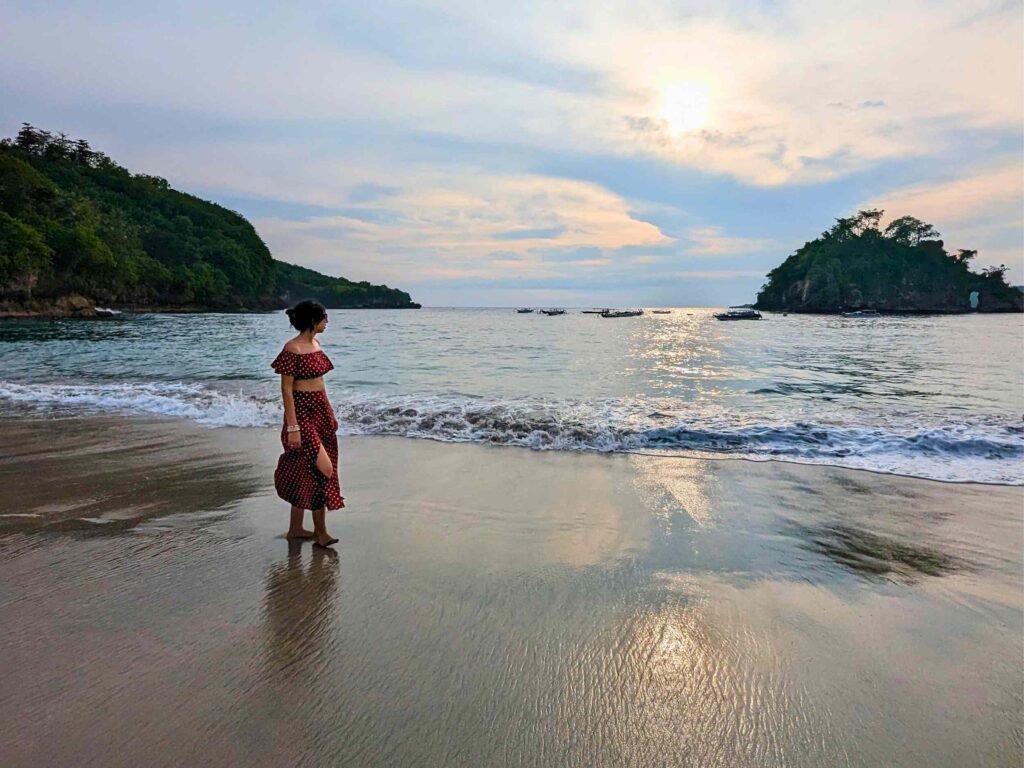 A serene beach scene at Crystal Bay, showing the tour leader standing barefoot at the water's edge on wet sand, looking out at the ocean. She is wearing a matching dark red polka-dot two-piece top and long skirt. In the background, the sun sets behind light clouds, casting a soft glow over the calm water, distant boats, and two lush, tree-covered rocky islands flanking the bay.