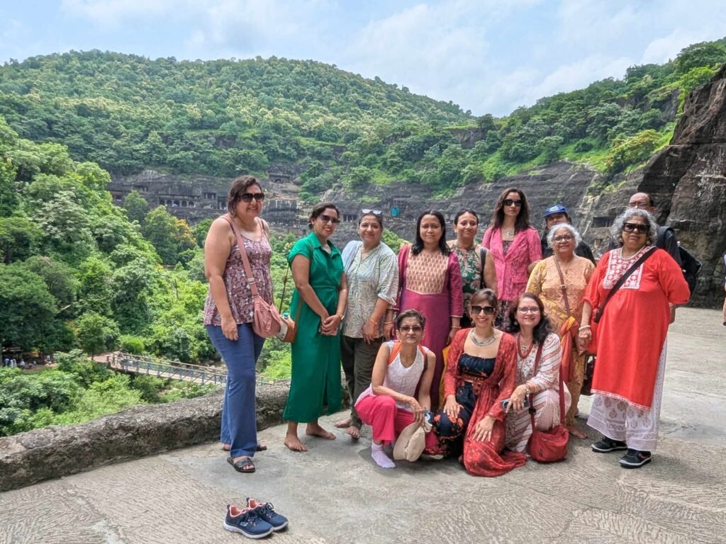 A group of people stand and pose for a photo on an elevated stone path, with a green, tree-covered mountain and ancient Ajanta caves facades visible across the gorge behind them.