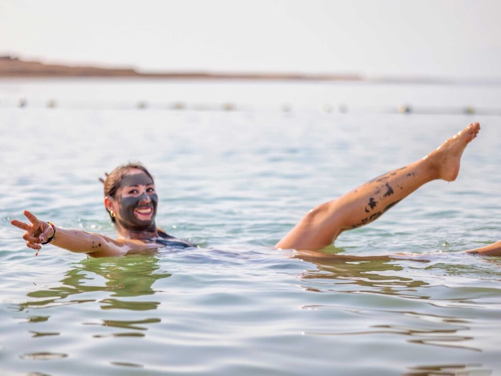A smiling woman with a mud mask on her face floating effortlessly in the Dead Sea while making a peace sign.