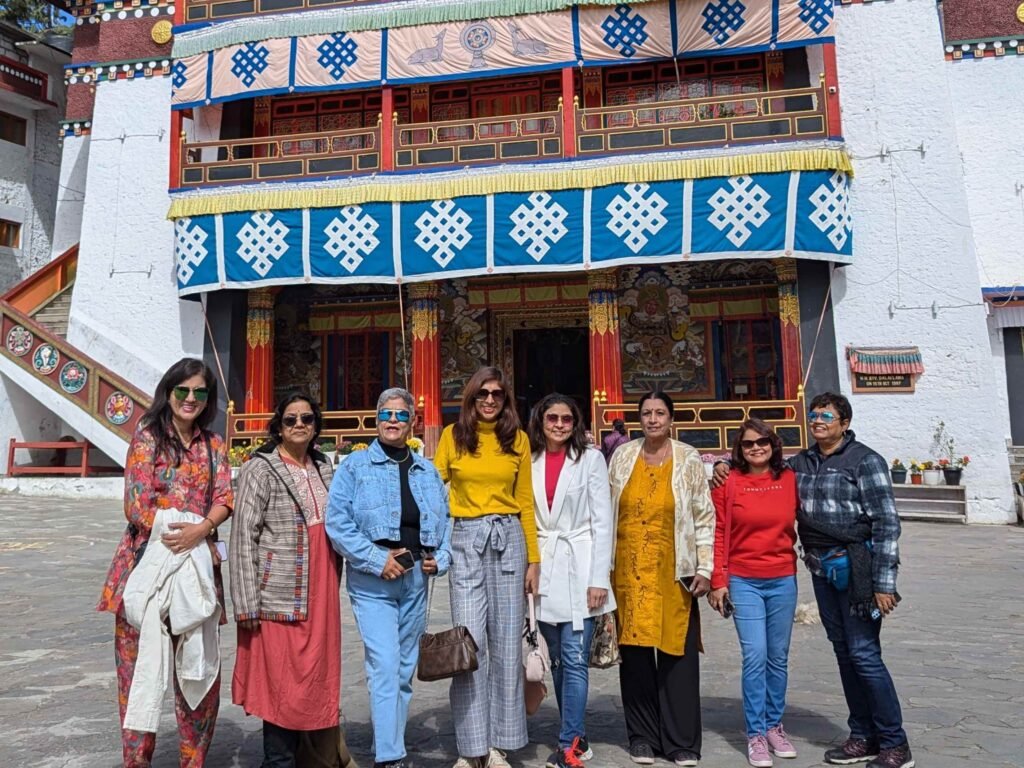 A group of eight women travelers posing together in front of the ornate, white-walled Tawang Monastery in Arunachal Pradesh, featuring traditional Tibetan architectural details, colorful murals, and blue banners with white "endless knot" symbols.