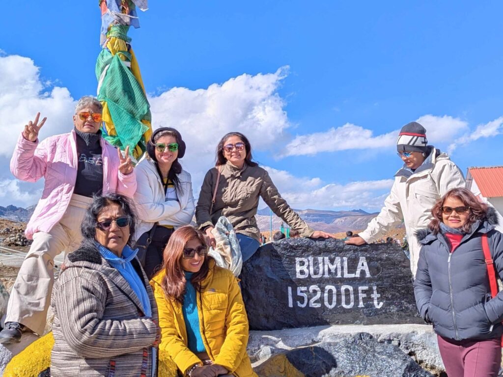 A group of six tourists in warm winter clothing posing beside a prominent black mountain marker inscribed with "BUMLA 15200FT" at the high-altitude Bum La Pass, with the expansive, rocky Himalayan landscape and a bright blue sky stretching into the distance.