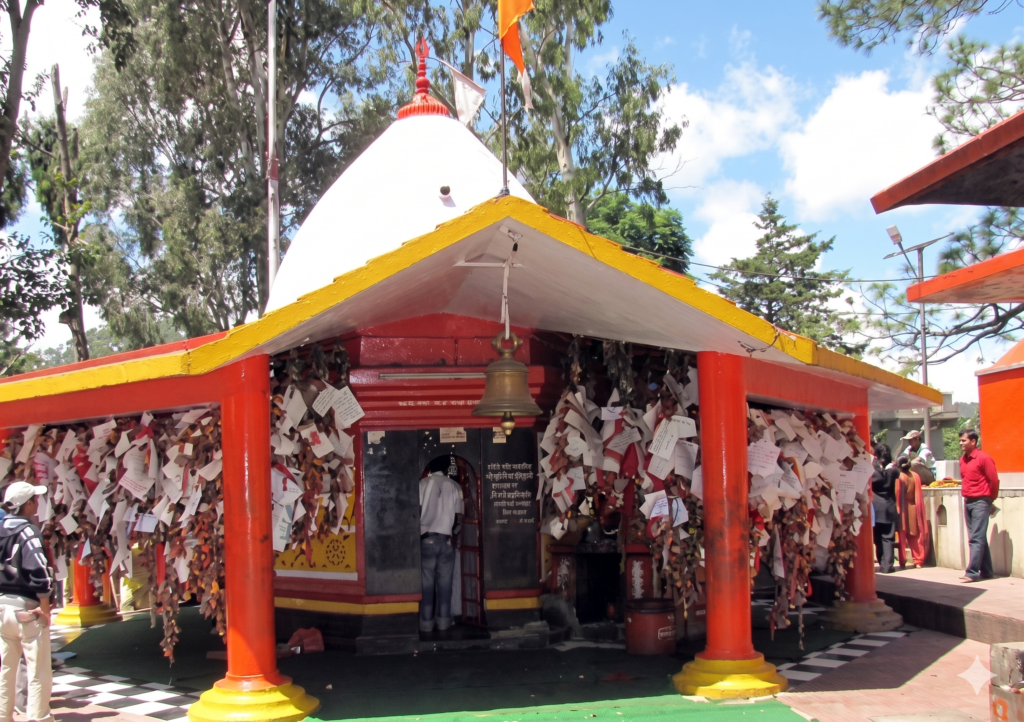 A vibrant shot of the Chitai Golu Devta Temple in Uttarakhand, featuring a white dome, red pillars, and yellow eaves from which countless white letters and bells are hung, with devotees nearby.