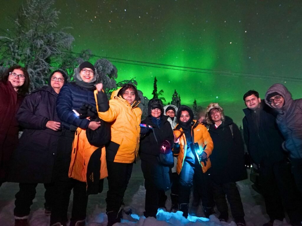 A group of about ten people wearing heavy winter coats, hoods, and hats stand together outside at night in the snow. They are smiling at the camera, with some holding small lights. In the background, a bright green aurora borealis glows in a starry night sky above snow-covered trees.