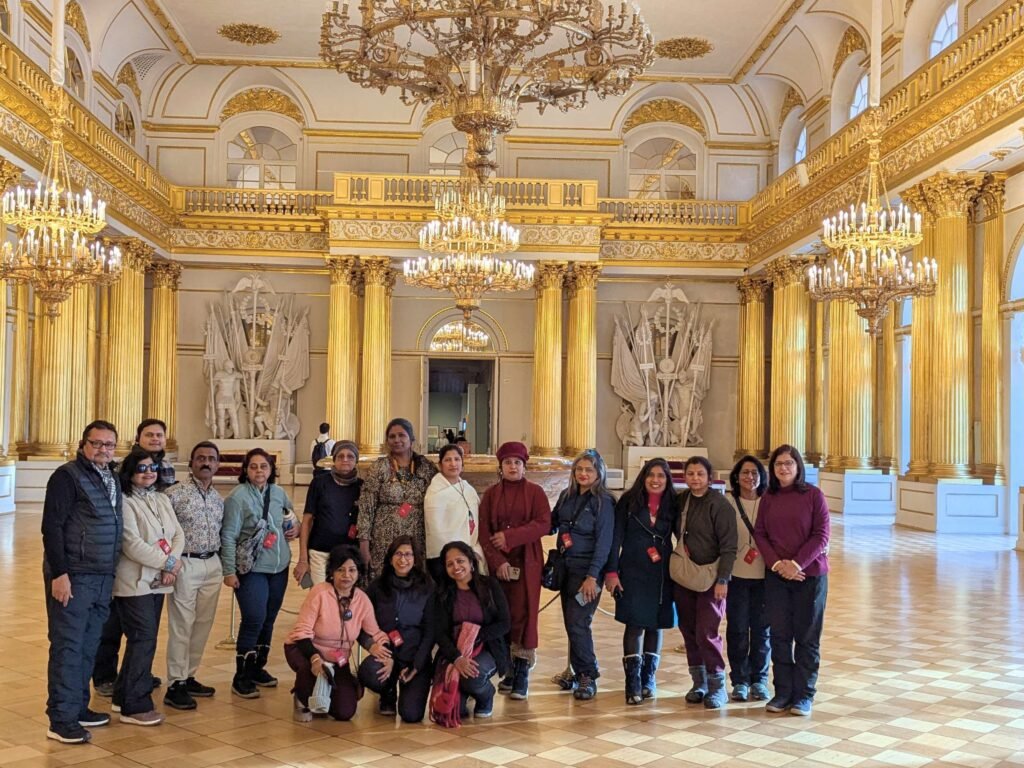 Group of tourists standing together in the ornate, golden Armorial Hall of the State Hermitage Museum in St. Petersburg beneath crystal chandeliers.