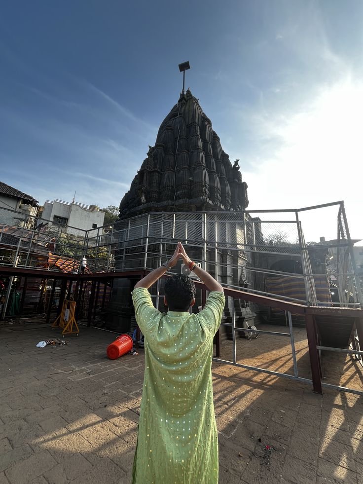 Rear view of a man in a green traditional kurta with hands raised in prayer before a dark stone Hindu temple spire under a blue sky