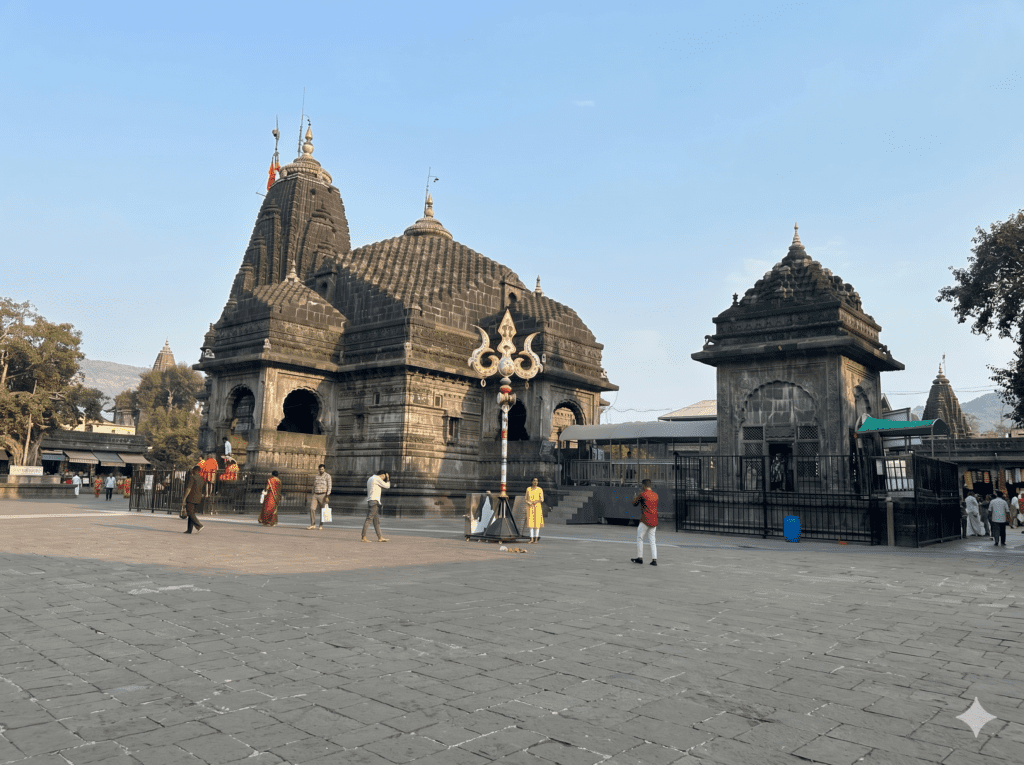 An exterior view of the ancient stone Tryambakeshwar Temple with a prominent decorative Trishul in the courtyard under a clear sky