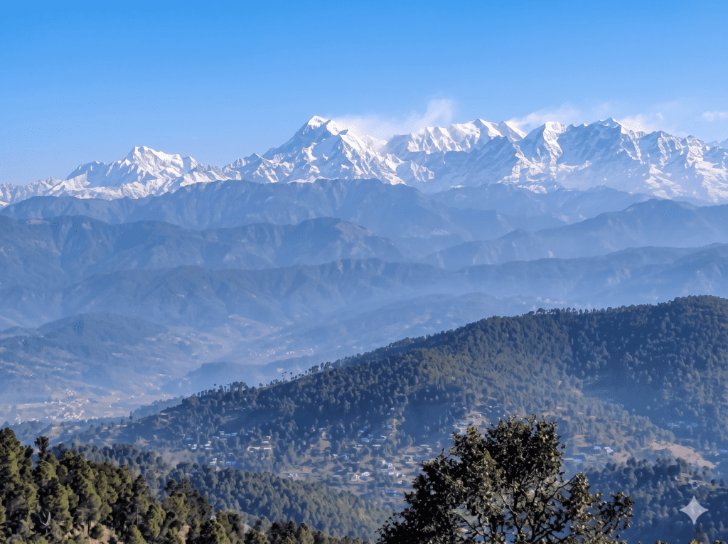 A wide landscape photograph capturing the Kausani mountain view. In the background, majestic snow-capped Himalayan peaks are sharply visible under a clear blue sky. Layers of hazy, forested foothills fill the mid-ground, featuring a small village and winding roads on the slopes. Dark green pine trees frame the lower foreground.
