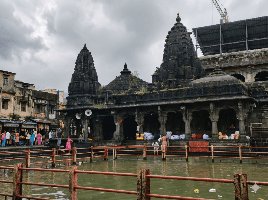 A historic, dark stone Hindu temple with intricate carvings and spires sits next to a green-water sacred bathing pool under an overcast sky