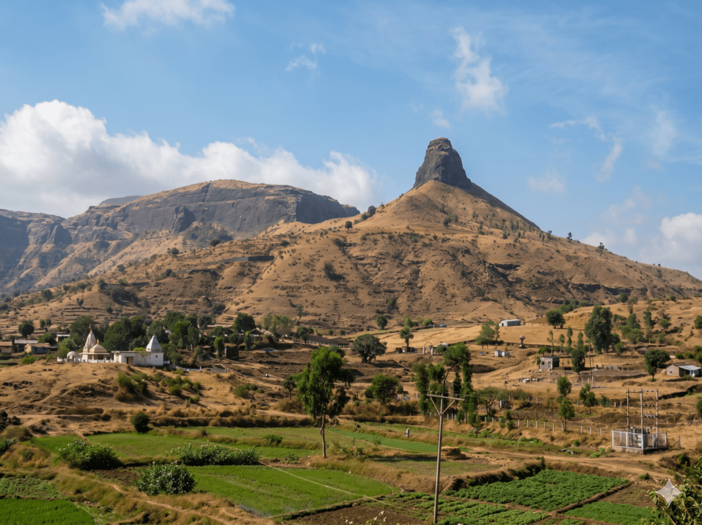 A prominent, steep rocky hill towering over a dry, grassy landscape with small structures and green fields at the base under a blue sky