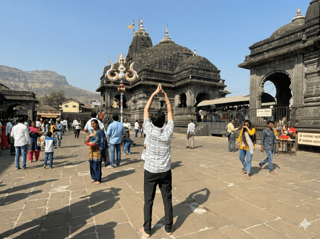 A person stands barefoot with arms raised in prayer facing the stone structure of Trimbakeshwar Temple