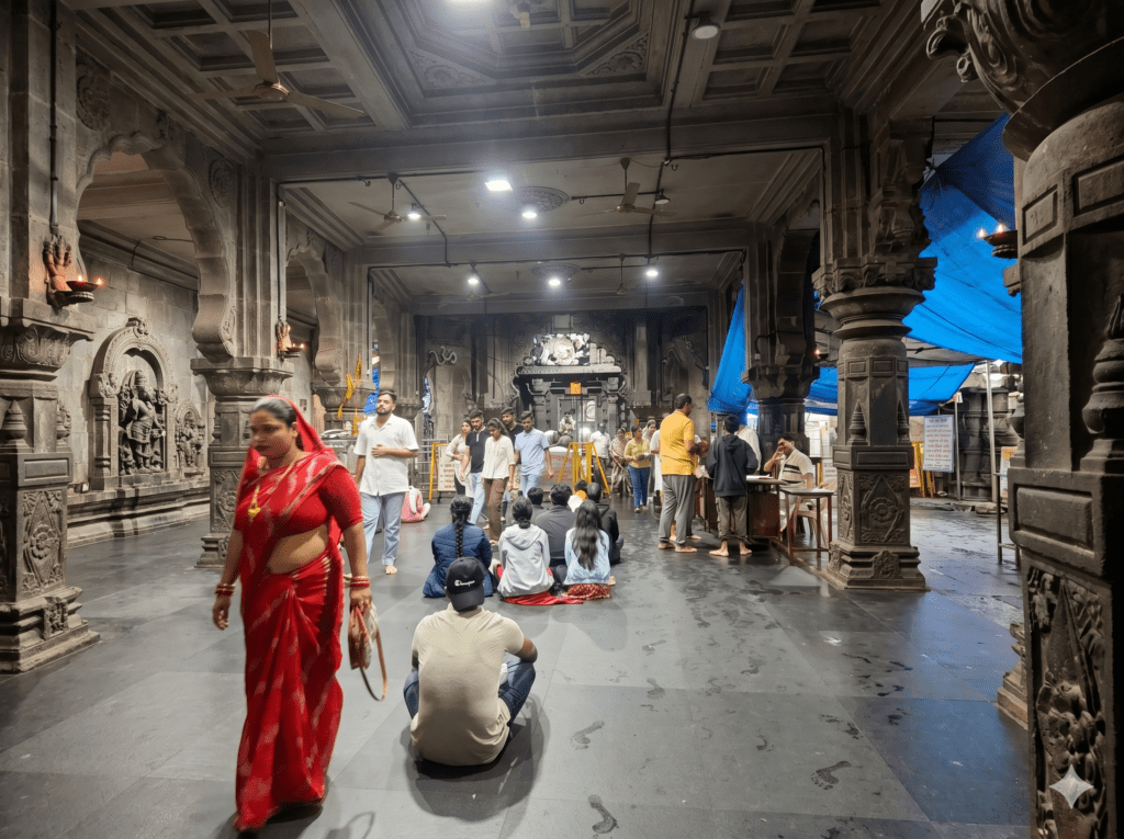 A wide view of the stone pillared interior of a Hindu temple hall with devotees waiting and walking