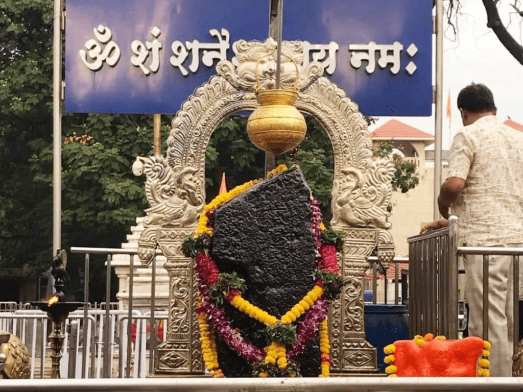 A large, black rock idol representing Lord Shani, adorned with yellow and maroon flower garlands. A golden pot hangs directly above the idol, which is framed by an ornate silver arch. A blue sign in the background reads "Om Sham Shanaishcharaya Namah" in Devnagari script.