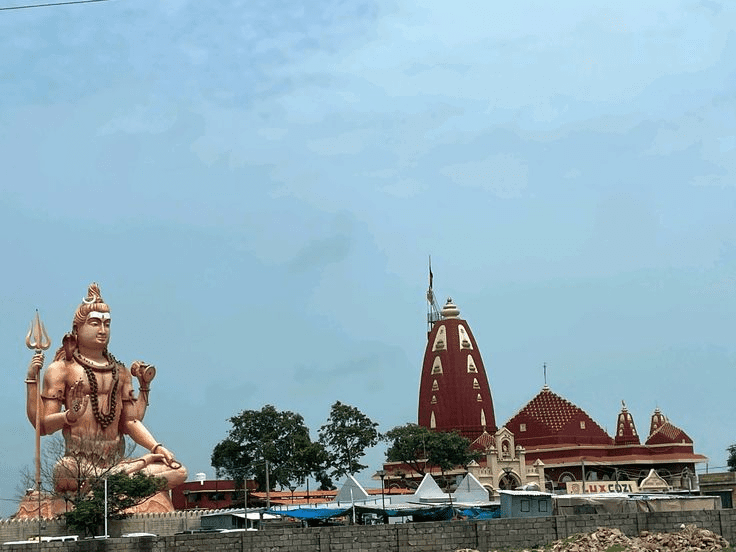 A massive orange statue of Lord Shiva sitting in meditation next to the red-spired Nageshwar Temple