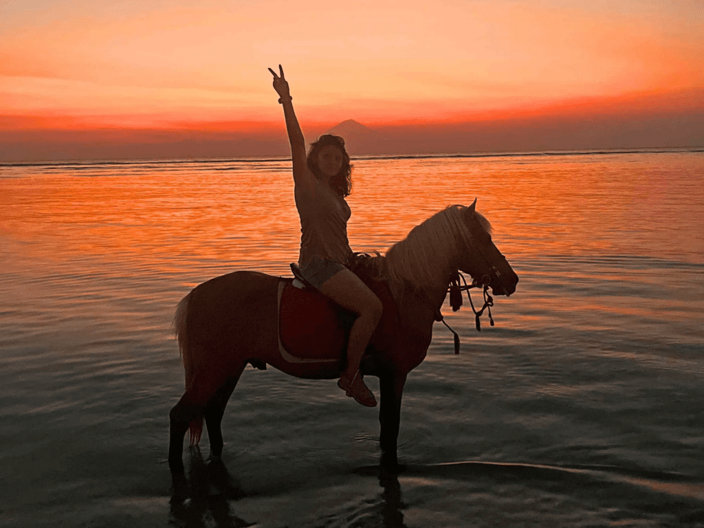 Silhouette of a woman riding a horse in the shallow ocean water during a vibrant orange sunset on Gili Trawangan island, with a volcano visible on the distant horizon.