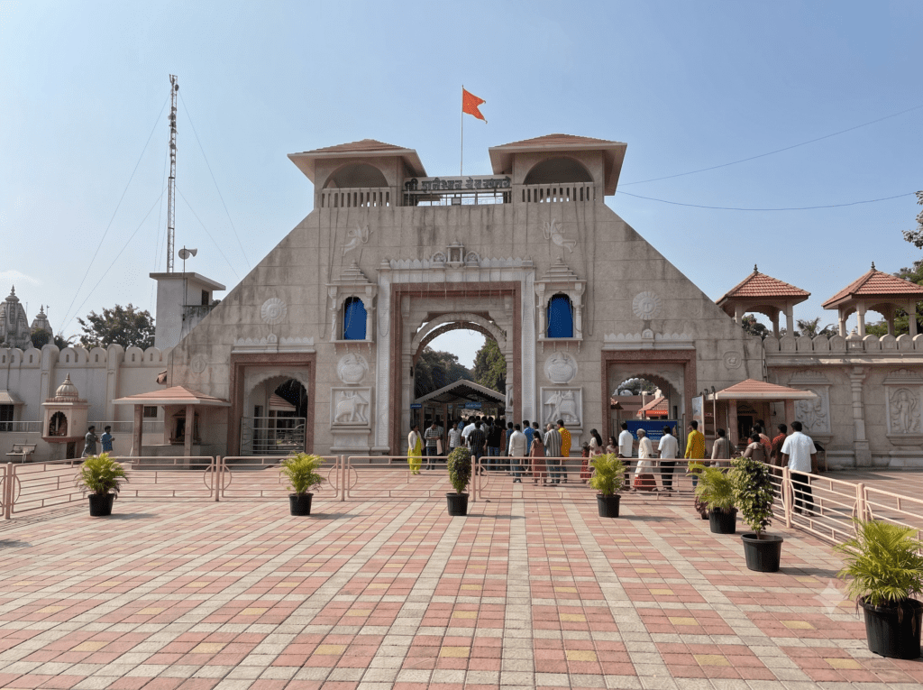 A wide-angle view of the large stone entrance gate of Shani Shingnapur temple with people walking through under a blue sky.
