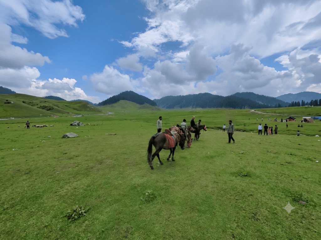 A wide-angle view of people riding horses on a vast green field in a mountain valley under a cloudy sky.