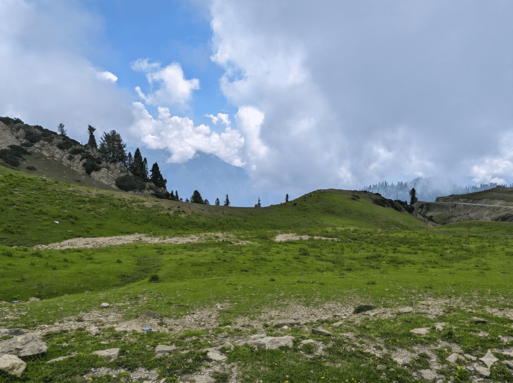 A panoramic roadside landscape showing a vibrant green mountain slope and rolling valley beneath heavy clouds near the Peer Baba Dargah.