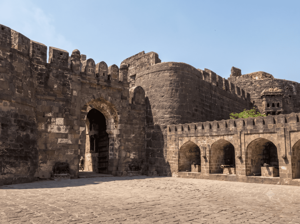 A large stone archway entrance and fortified walls of Daulatabad Fort with small alcoves holding old cannons on the right.