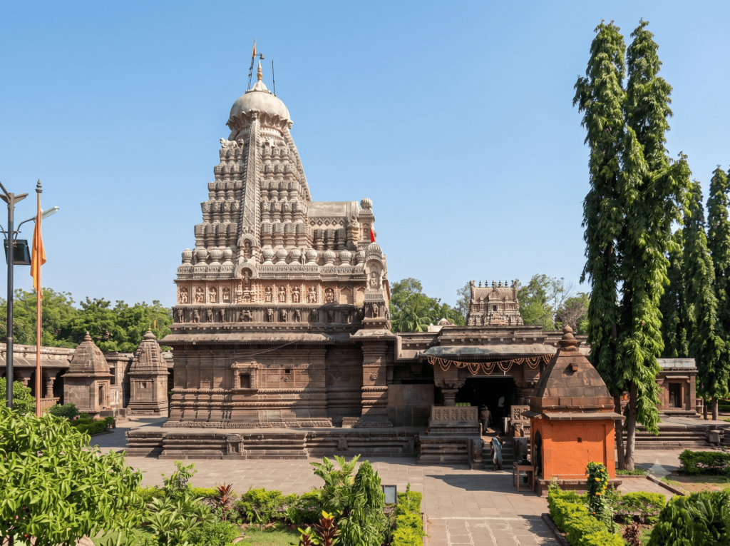 A wide shot of the stone Grishneshwar Jyotirlinga Temple with an intricately carved shikhara tower, surrounded by a stone courtyard and green shrubbery.