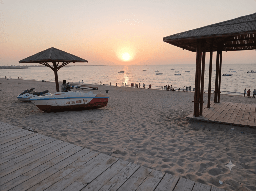 A scenic sunset over Shivrajpur Beach with people, boats, and a wooden walkway.