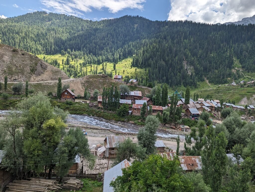 High-angle view of a village in Tulail Valley with wooden houses next to a rushing river, backed by a massive, pine-covered mountain under a blue, cloudy sky.
