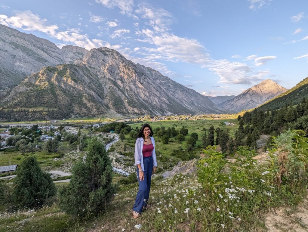 A smiling woman standing amidst white wildflowers on Khandiyal Top, looking out over a vast green valley and majestic mountain ranges in Gurez.