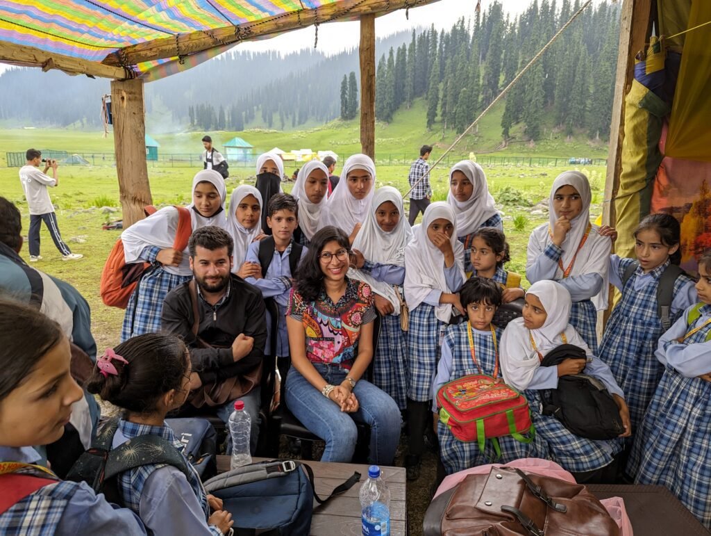 A large group of students in blue plaid school uniforms and white hijabs gather around a man and a woman under a wooden shelter, with a green valley and pine trees in the background.