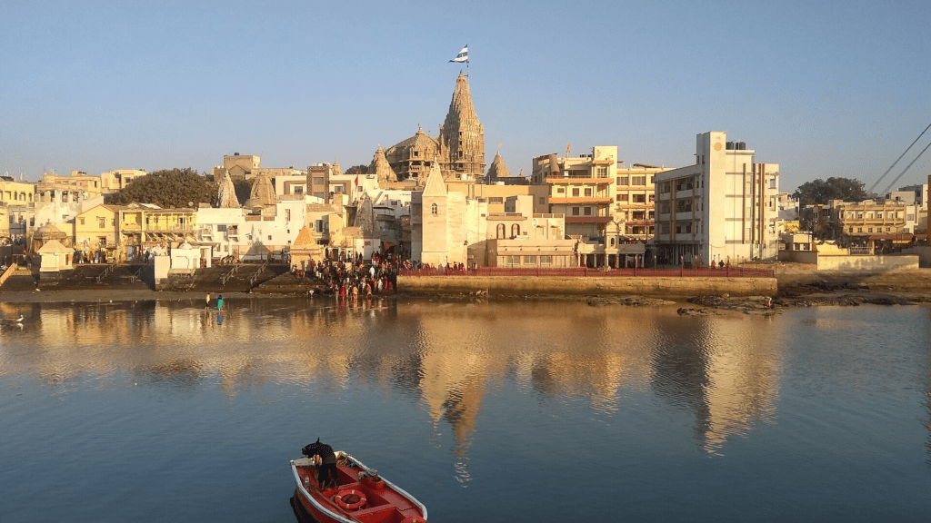 Scenic view of Dwarkadhish Temple and town buildings reflecting on the Gomti River with a red boat in the foreground.