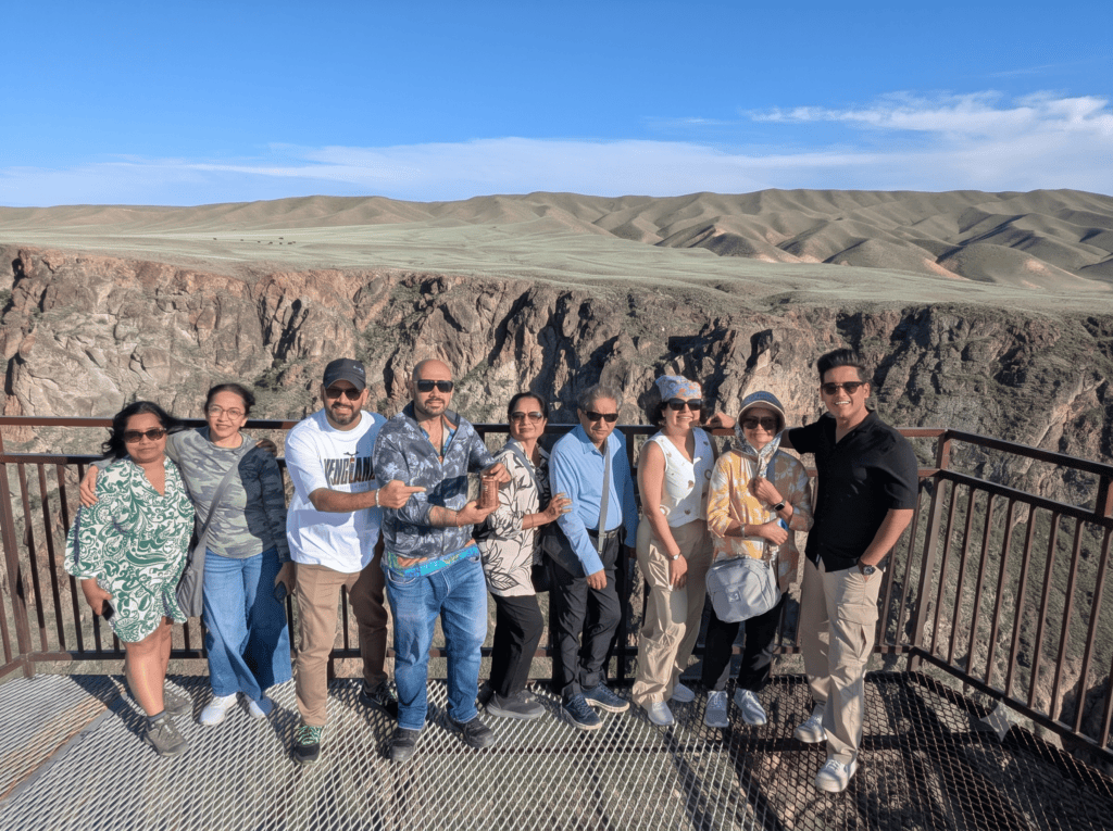 A group of nine tourists posing together on a metallic lookout platform overlooking a deep canyon.