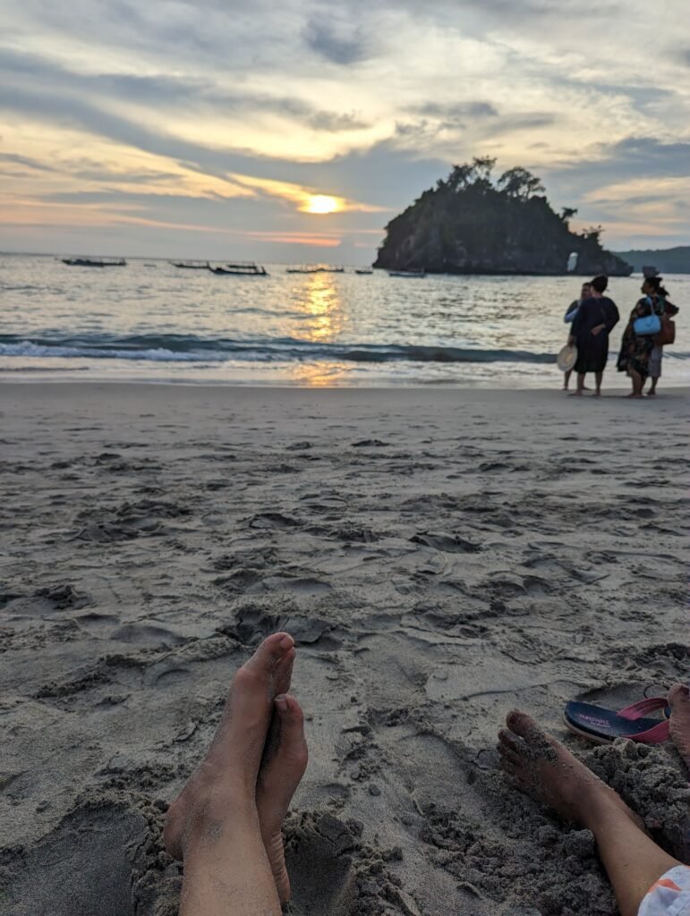 First-person view of feet resting on a sandy beach during a dramatic sunset, with a rock arch formation in the ocean.