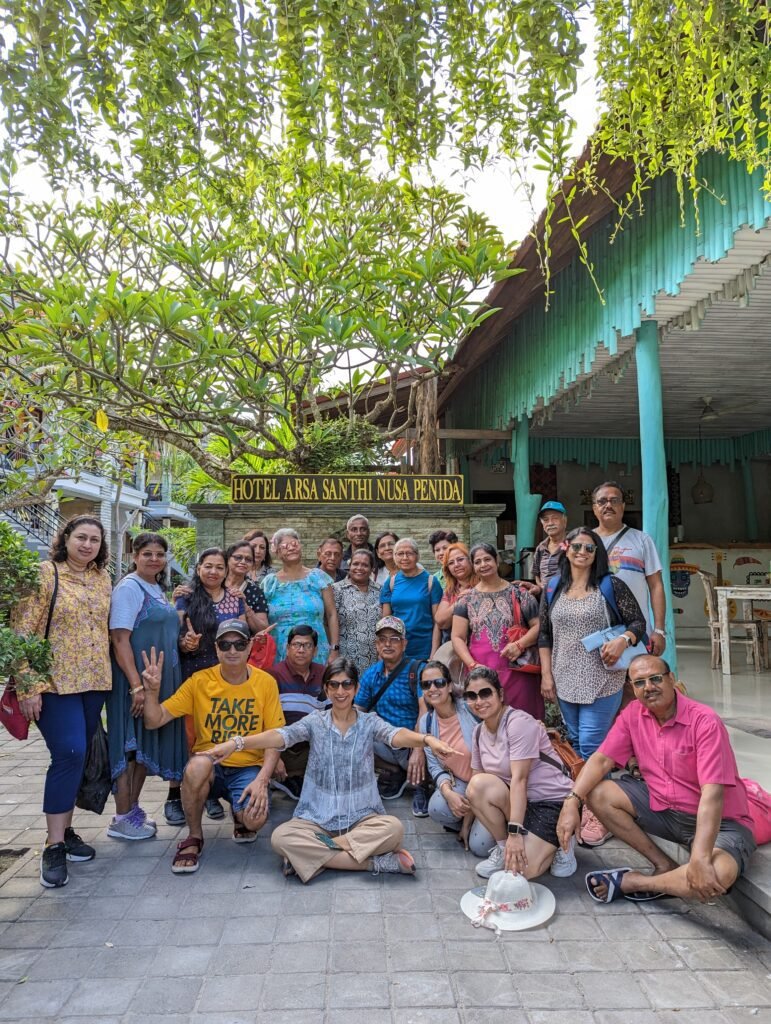 A diverse group of about twenty people smiling and posing in a courtyard beneath a large tree and a hotel sign.