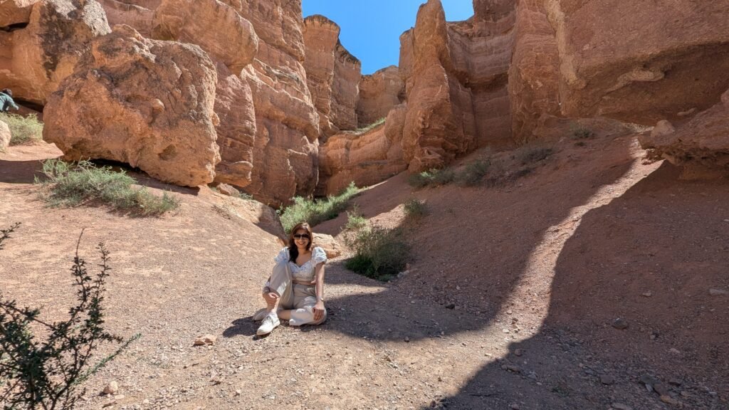 Woman in sunglasses sitting on dirt ground between steep red canyon cliffs.