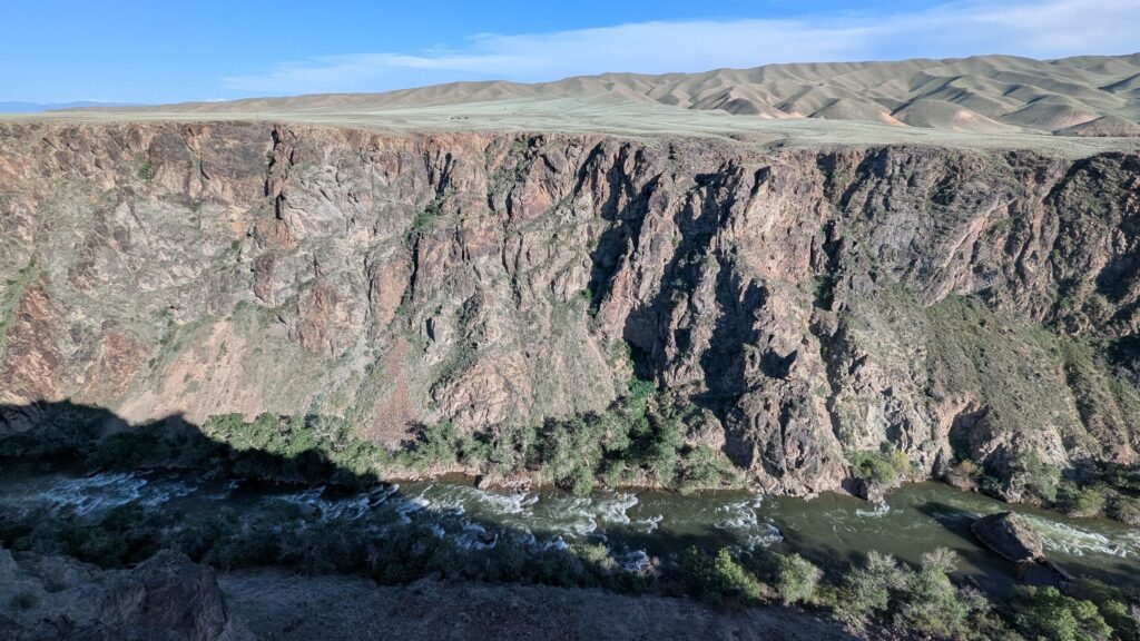 Rocky canyon cliff looking down at a rushing river with green hills in the distance.