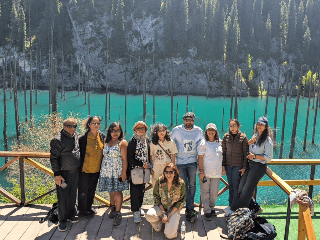 A travel group posing together on a wooden boardwalk overlooking the turquoise waters and sunken trees of Lake Kaindy in Kazakhstan.