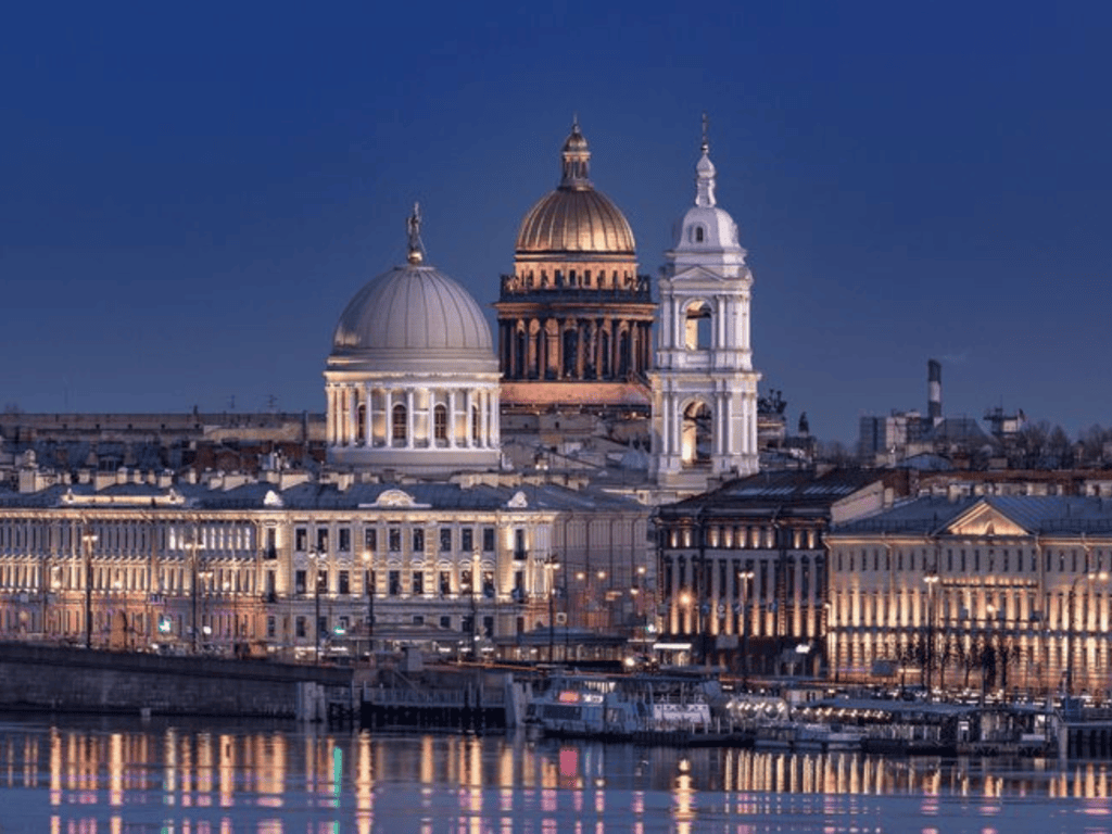 Night view of Saint Petersburg's illuminated skyline and riverfront buildings featuring the golden dome of Saint Isaac's Cathedral reflected in the Neva River.