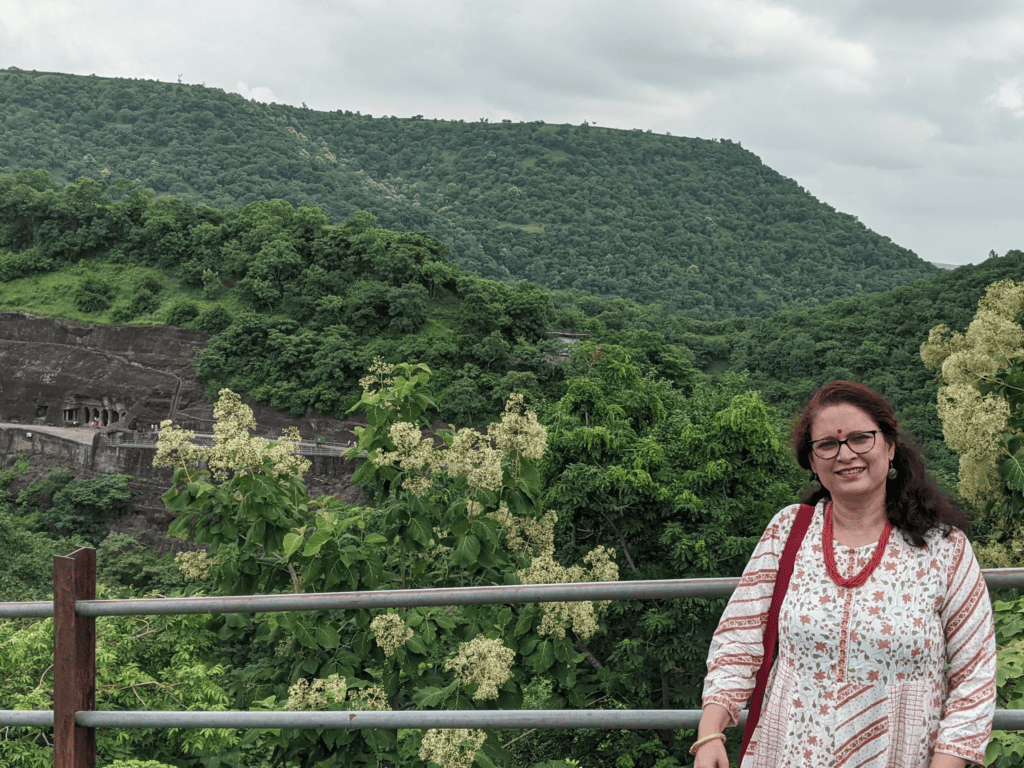 A smiling woman with glasses and traditional red-and-white patterned attire standing outdoors in front of a metal railing, with lush green hills and the rock-cut Ajanta Caves visible across the valley in the background.