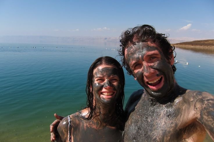 A smiling man and woman with dark Dead Sea mud smeared over their faces and bodies standing by the water.