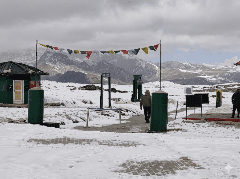 A photograph of colorful Tibetan prayer flags strung across a metal archway at a snowy mountain pass checkpoint, with a person walking through and snow-capped peaks in the background under a cloudy sky.