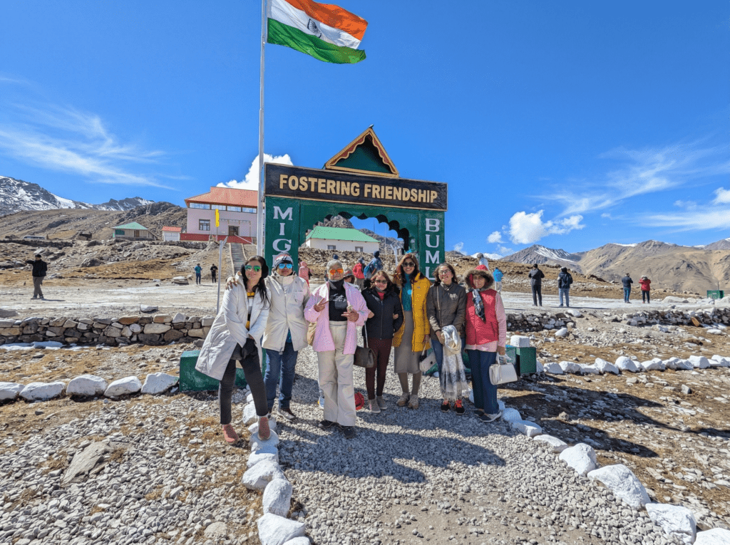 A group of travelers in heavy winter jackets posing under the 'Fostering Friendship' sign at Bumla Pass, Arunachal Pradesh, showcasing the necessary warm attire for the high-altitude border.