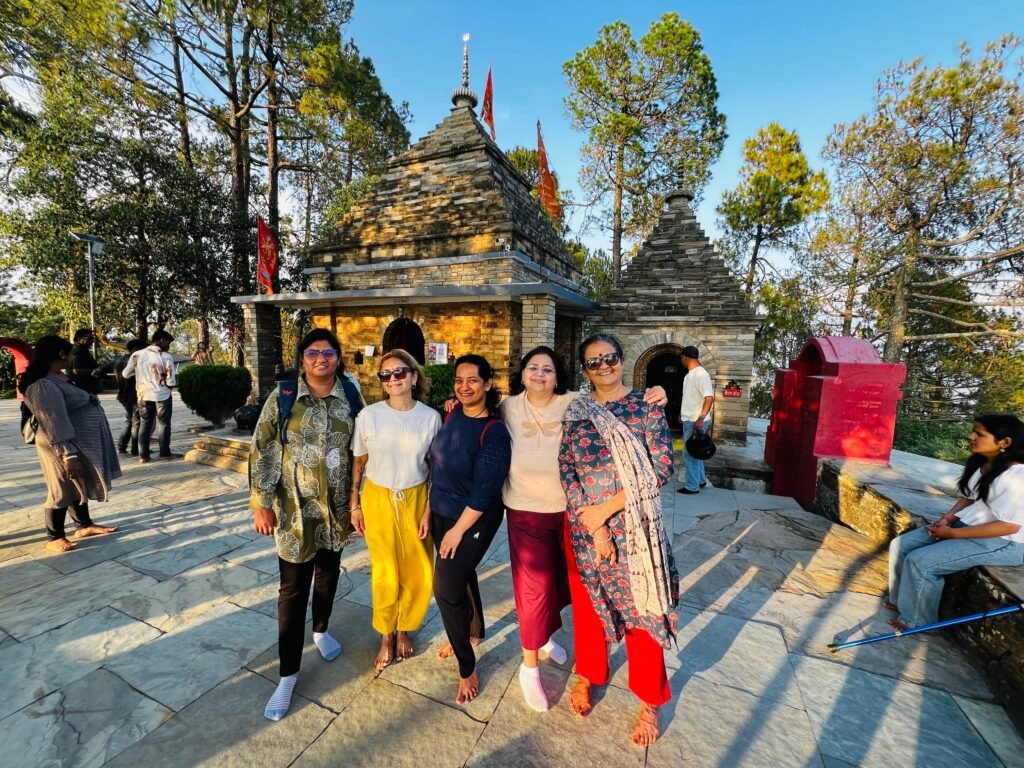 A group of five women smiling and posing at Kasar Devi Temple on a stone path, with ancient stone temple structures and trees under a blue sky.
