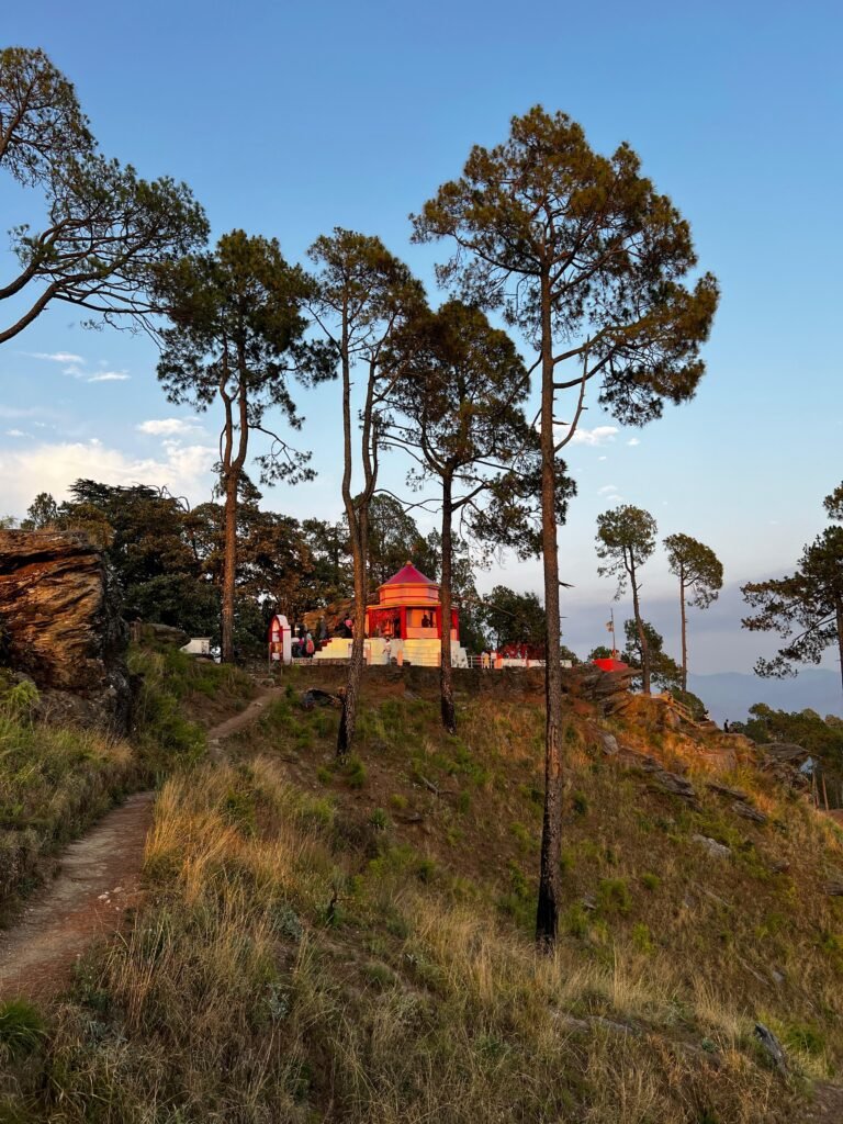 A vibrant red and white Kasar Devi Temple stands atop a grassy hill surrounded by tall pine trees under a clear blue sky.