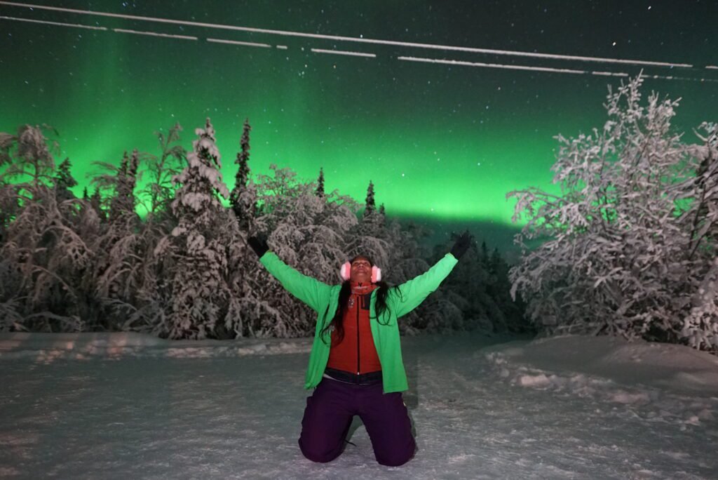 A person kneeling in the snow with arms raised in joy beneath a vivid green aurora borealis in the night sky.