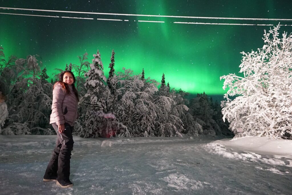 Nighttime astrophotography portrait of a person in a snow-covered forest with a long exposure of green northern lights in the sky.