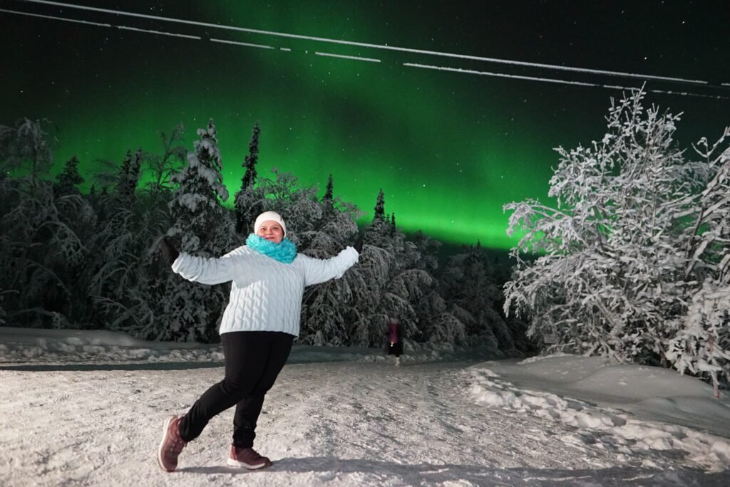 Woman wearing a knit sweater, fleece infinity scarf, beanie, dark thermal pants, and gloves standing in deep snow under the aurora.