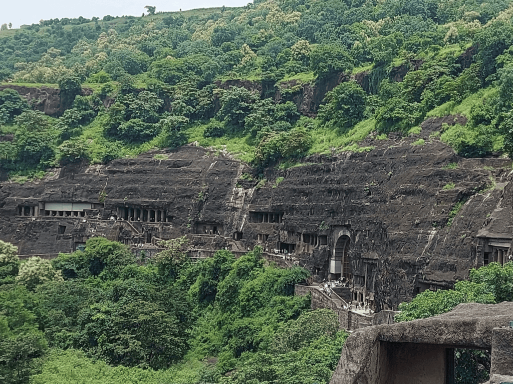 A panoramic view of a massive, rocky cliffside featuring multiple ancient cave entrances with pillars and walkways, surrounded by green trees.